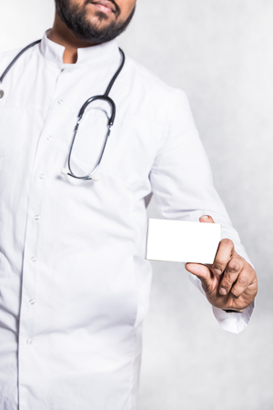 Handsome young doctor in a white coat with a stethoscope holds on the outstretched hand a blank white package box for blister of pills. Mock up.の写真素材