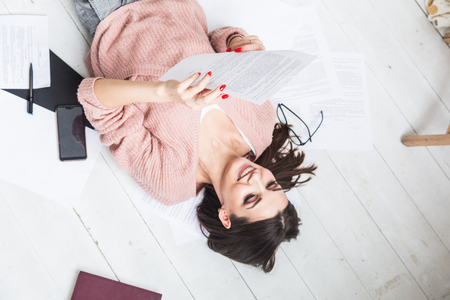 A beautiful woman lies on the floor among papers and documents, the girl freelancer smiles and relaxes during a break from workの写真素材