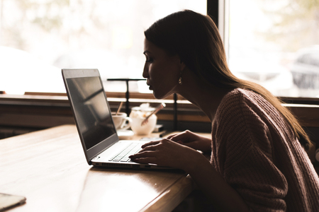 Attractive young woman sits at a table in a cafe with a cup of coffee and enjoys a laptopの写真素材