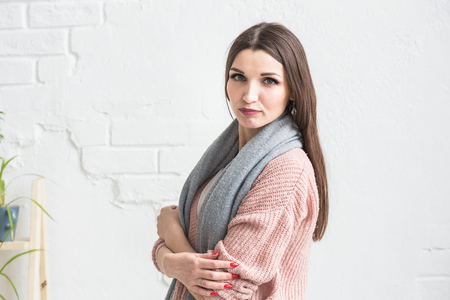 Portrait of a beautiful young brunette woman in a sweater with a scarf on a light background.の写真素材
