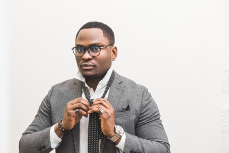 Young African American businessman in a gray suit tying a tie on a white background.の写真素材
