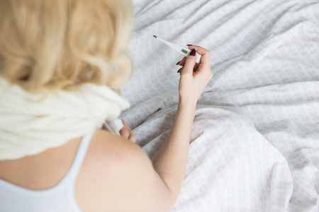Beautiful blond woman sitting in bed with a thermometer and a scarf wrapped around the neck and a handkerchief. illness and health at homeの写真素材