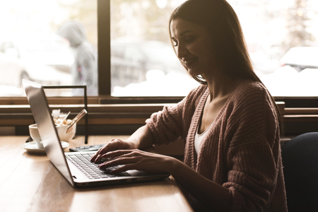 Attractive young woman sits at a table in a cafe with a cup of coffee and enjoys a laptopの写真素材