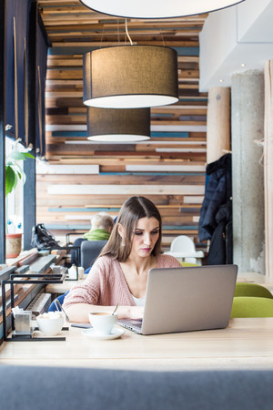 Attractive young woman sits at a table in a cafe with a cup of coffee and enjoys a laptopの写真素材