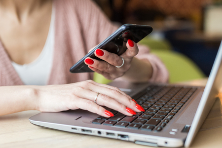 Attractive young woman sits at a table in a cafe with a cup of coffee and enjoys a laptop and smartphoneの写真素材