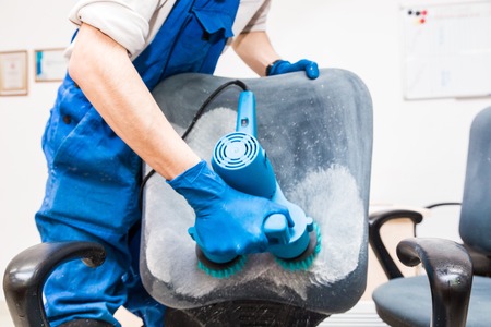 Young man in workwear and rubber gloves cleans the office chair with professional equipment.の写真素材