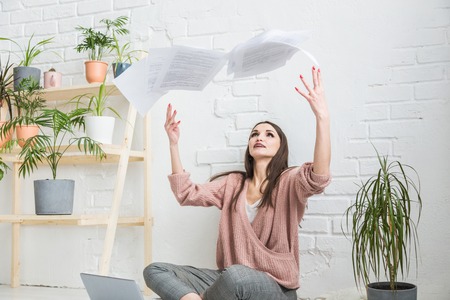A young happy woman throws up a bunch of pieces of paper and documents sitting near the laptop, the...