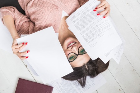 A beautiful woman in stylish glasses lies on the floor among the papers and documents, the girl freelancer smiles and relaxes during a break from workの写真素材