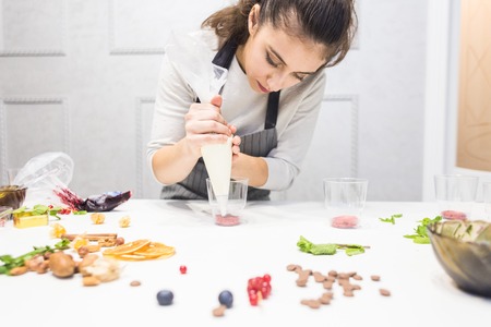 A confectioner prepares a trifle in three cups. Desserts are on the white table in the kitchen. The concept of homemade pastry, cooking cakes.の写真素材