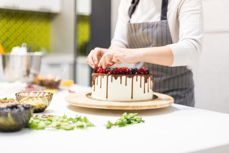 Confectioner decorates with berries a biscuit cake with white cream and chocolate. Cake stands on a wooden stand on a white table. The concept of homemade pastry, cooking cakes.の写真素材