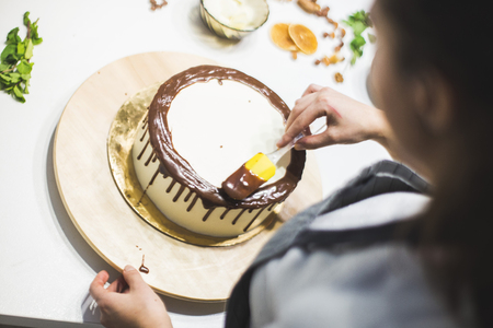 The confectioner decorates liquid chocolate biscuit cake with white cream, standing on a wooden stand. The concept of homemade pastry, cooking cakes.の写真素材