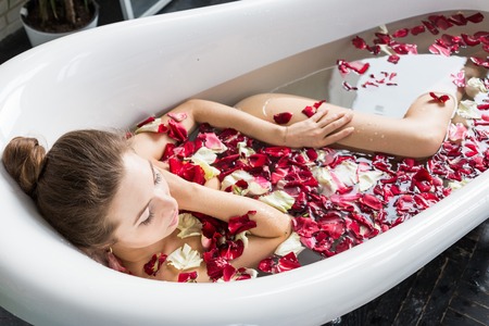 A young attractive girl takes a bathroom with flower petals and relaxes against the background of a beautiful light interior. Spa treatments for beauty and health with skin careの写真素材