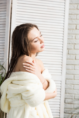 Attractive young girl in a white bathrobe applies cream to the skin while sitting in the interior of the bathroom and doing morning procedures. Skin and body careの写真素材