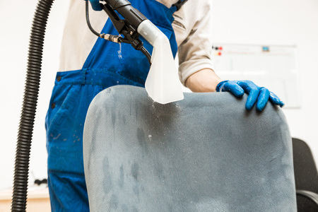 Young man in workwear and rubber gloves cleans the office chair with professional equipment.