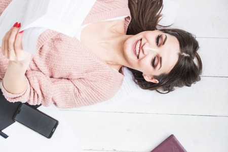A beautiful woman lies on the floor among papers and documents, the girl freelancer smiles and relaxes during a break from workの写真素材