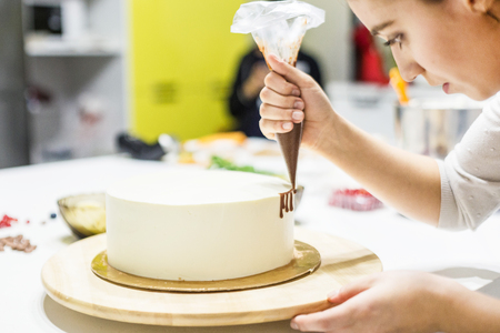 A confectioner squeezes liquid chocolate from a pastry bag onto a white cream biscuit cake on a wooden stand. The concept of homemade pastry, cooking cakes.の写真素材