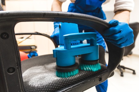 Young man in workwear and rubber gloves cleans the office chair with professional equipment.の写真素材
