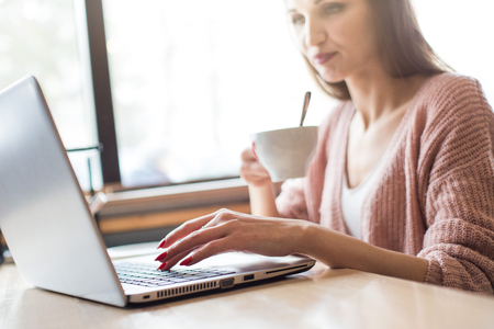 Attractive young woman sits at a table in a cafe with a cup of coffee and enjoys a laptopの写真素材