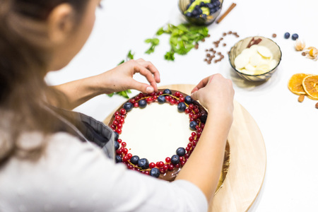 Confectioner decorates with berries a biscuit cake with white cream and chocolate. Cake stands on a wooden stand on a white table. The concept of homemade pastry, cooking cakesの写真素材