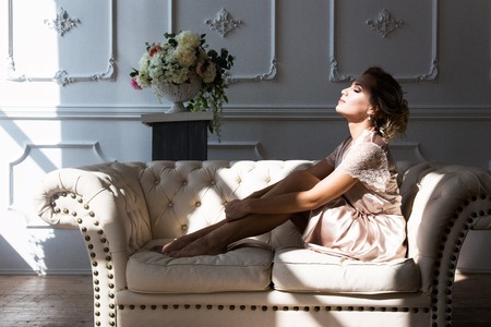 Beautiful young sexy blonde woman in a silk robe sitting on a couch hugging her legs in front of a...