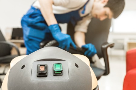 Young man in workwear and rubber gloves cleans the office chair with professional equipment.の写真素材