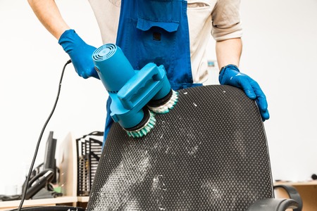 Young man in workwear and rubber gloves cleans the office chair with professional equipment.の写真素材