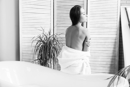 Attractive young girl in a white bathrobe applies cream to the skin while sitting in the interior of the bathroom and doing morning procedures. Skin and body careの写真素材