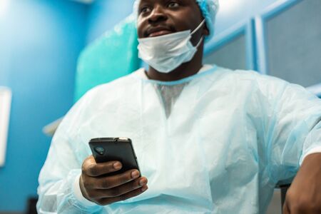 Portrait of a surgeon in uniform and medical mask after the operation is completed sitting with a phone in his hands.の写真素材