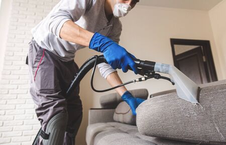 Cleaning service. Man janitor in gloves and uniform vacuum clean sofa with professional equipment