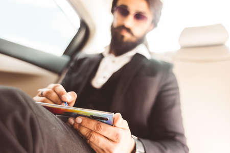 Stylish handsome Hindu businessman with glasses and a beard works in the backseat of the car and uses the phone. Safe and comfortable luxury travelの写真素材