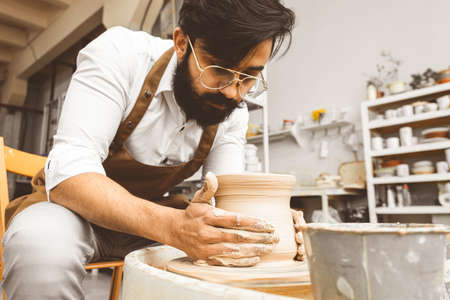 A young male potter is engaged in craft in his workshop on a potters wheel and makes a clay productの写真素材
