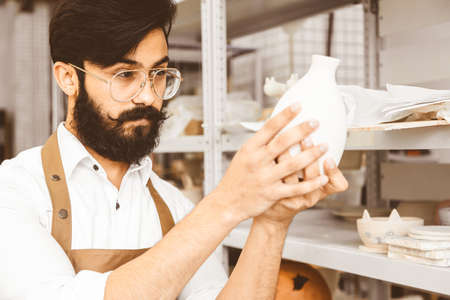 A young attractive male potter with a beard and mustache works in his workshop. Holds and inspects...