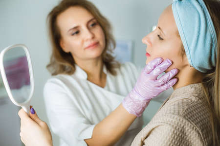Facial skin care and protection. A young woman at a beauticians appointment looks in a mirror. A specialist examines the skinの写真素材