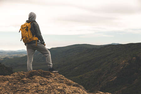 A man in hiking clothes with a yellow backpack stands on the top of a hill among the mountains...