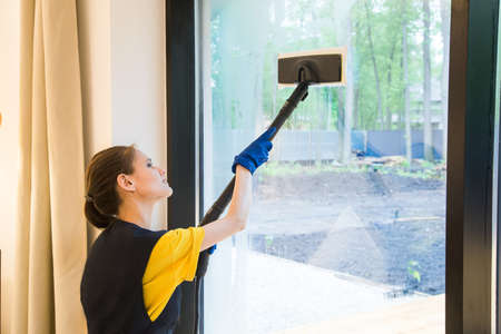 professional cleaning service. woman in uniform and gloves sponge washes panoramic windows in the cottage. washing steam mop and scraperの写真素材