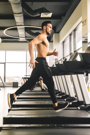 Handsome young sporty male bodybuilder with naked torso doing exercise on the treadmill in the gym