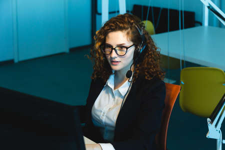 Portrait of a beautiful young woman operator with red curly hair and glasses in the interior of the...
