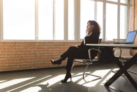 Portrait of a beautiful successful young business woman with red curly hair in an office interior sitting at a table and talking on the phoneの写真素材