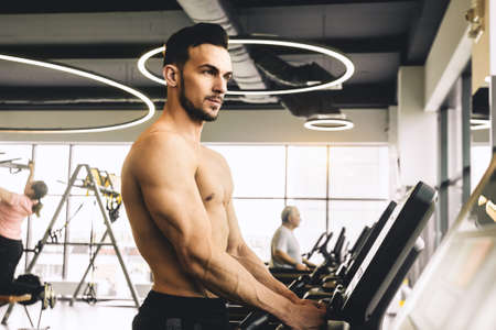 Handsome young sporty male bodybuilder with naked torso doing exercise on the treadmill in the gym