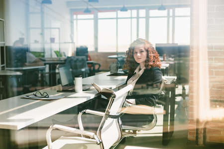 Portrait of a beautiful young business woman with red curly hair taking glass. The girl reads a book in the office. Successful career and leisure time and hobbiesの写真素材