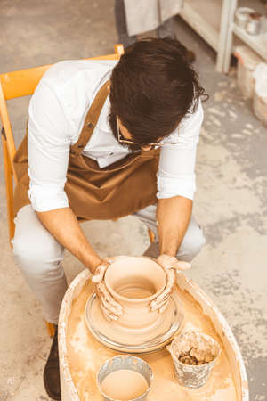 A young male potter is engaged in craft in his workshop on a potters wheel and makes a clay productの写真素材