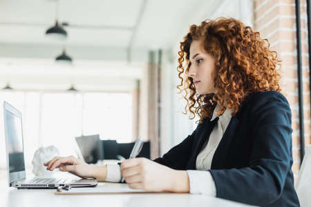 Beautiful young red-haired woman in a business suit works in an office with a laptop
