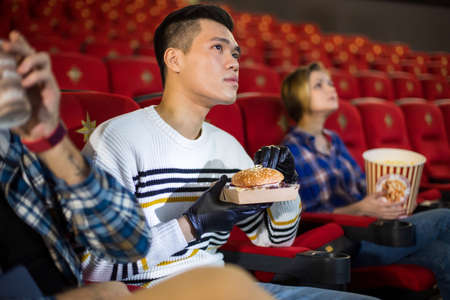 Friends watching a movie in the cinema and eat burgers. People sit in the armchairs of the cinema...