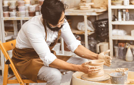 A young male potter is engaged in craft in his workshop on a potters wheel and makes a clay productの写真素材