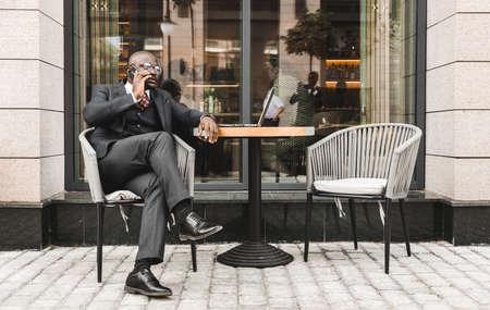 Portrait of a black African American businessman in a suit sitting in a city cafe outdoors and talking on the phone.の写真素材
