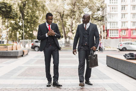 Two dark-skinned african american businessmen in suits and glasses walk outdoors in the city and...