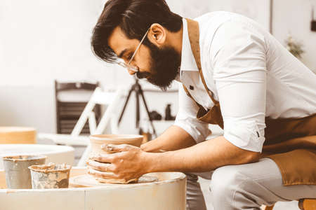 A young male potter is engaged in craft in his workshop on a potters wheel and makes a clay productの写真素材