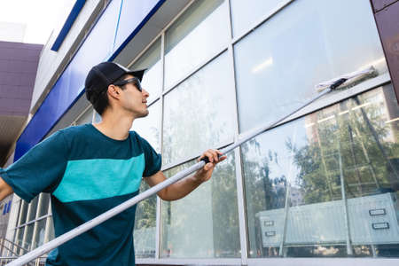 A young male worker of a professional cleaning service in overalls washes the glass of the windows...