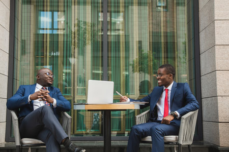 Two dark-skinned African American businessmen in suits and glasses with briefcases sit at a table in an outdoor city cafe.の写真素材