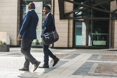 Two dark-skinned african american businessmen in suits and glasses walk outdoors in the city and discuss businessの写真素材
